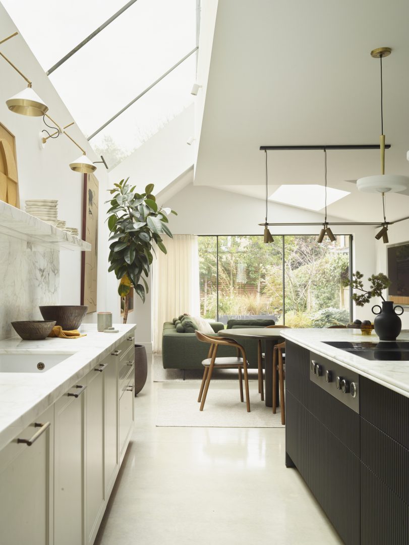 Bright bespoke kitchen with white cabinetry, dark fluted island, brass pendant lighting and large roof glazing overlooking an open-plan dining area.