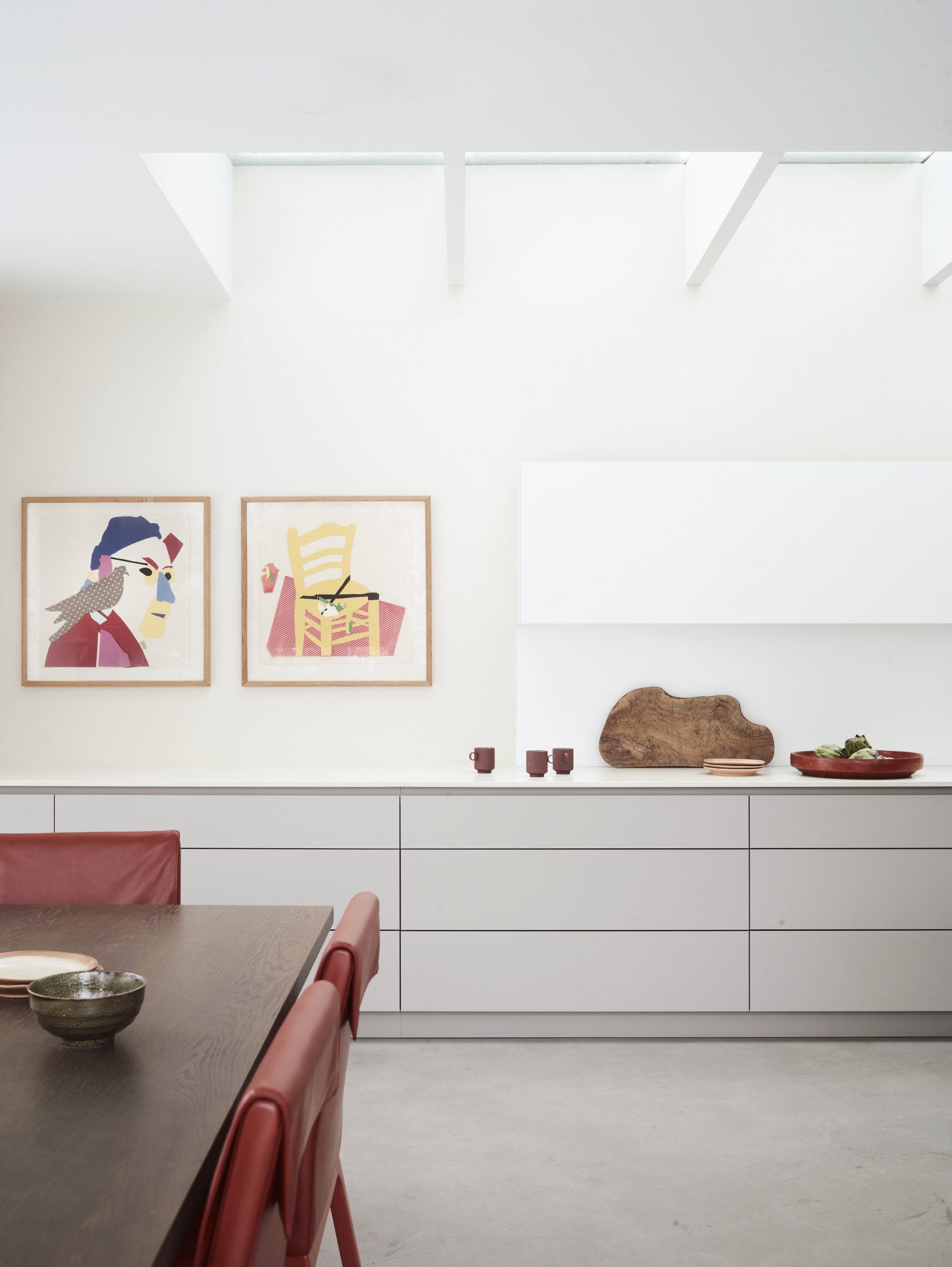 Minimalist white bespoke kitchen with handleless cabinetry, a sleek integrated extractor, contemporary artwork and a dining area with red leather chairs beneath an architectural skylight.