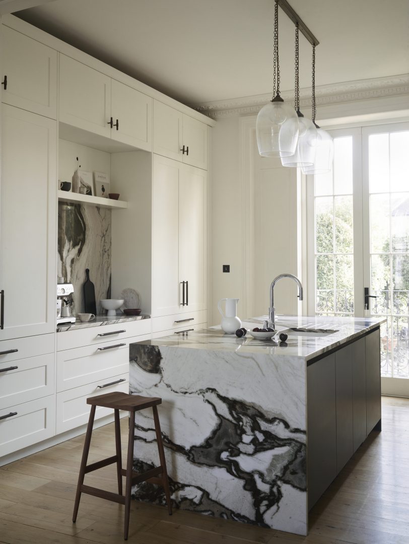 Luxury bespoke kitchen with white framed cabinetry, dramatic natural stone waterfall island and glass pendant lighting.