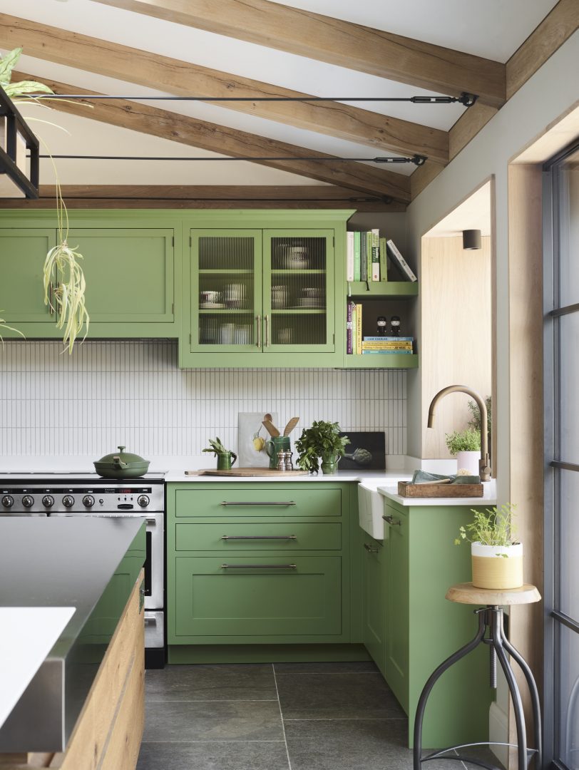 Green Shaker kitchen cabinetry with glazed wall cabinets, brass hardware, timber beams and farmhouse sink.