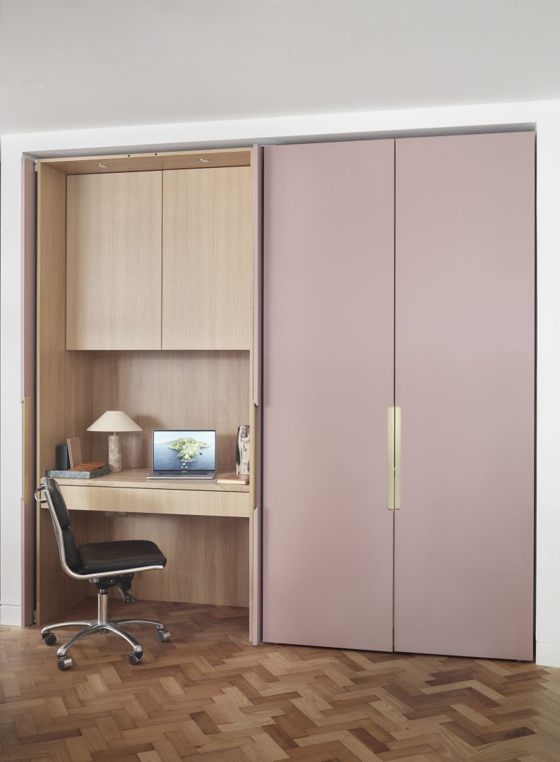 Concealed home office hidden behind bespoke pink cabinetry, with pocket doors revealing an integrated workspace within a seamless interior. To the right is a hidden coffee station behind pocket doors.