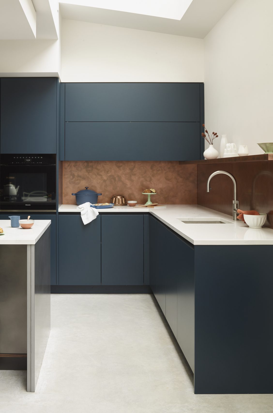Dark blue bespoke kitchen with Hague Blue cabinetry, antique copper splashback and light stone worktops alongside a patinated steel island.