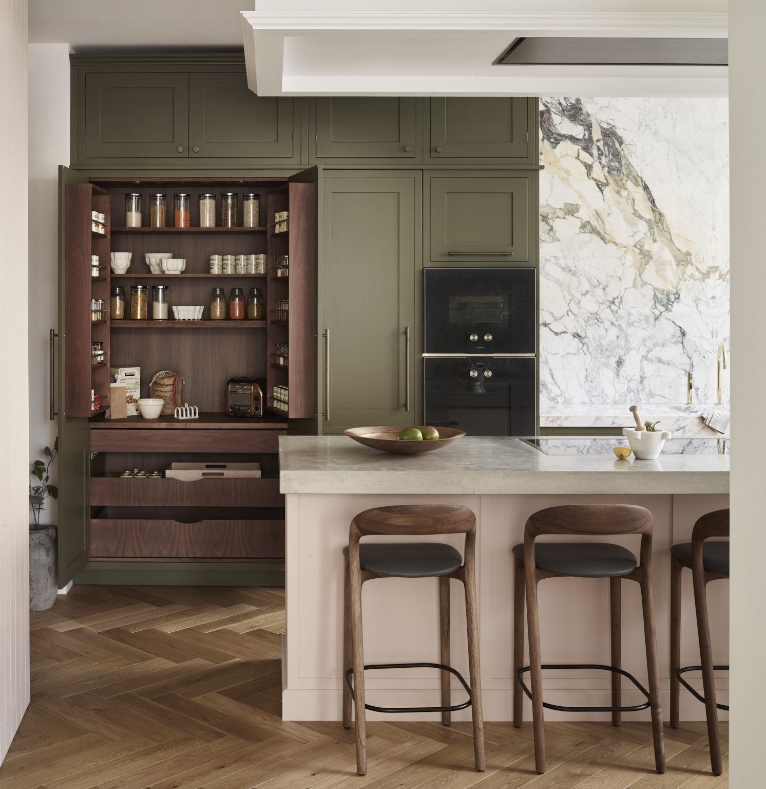 Classic green bespoke kitchen with a concealed walnut pantry. The contrasting soft pink island and poured concrete worktop are unified by the natural tones in the statement marble splashback.
