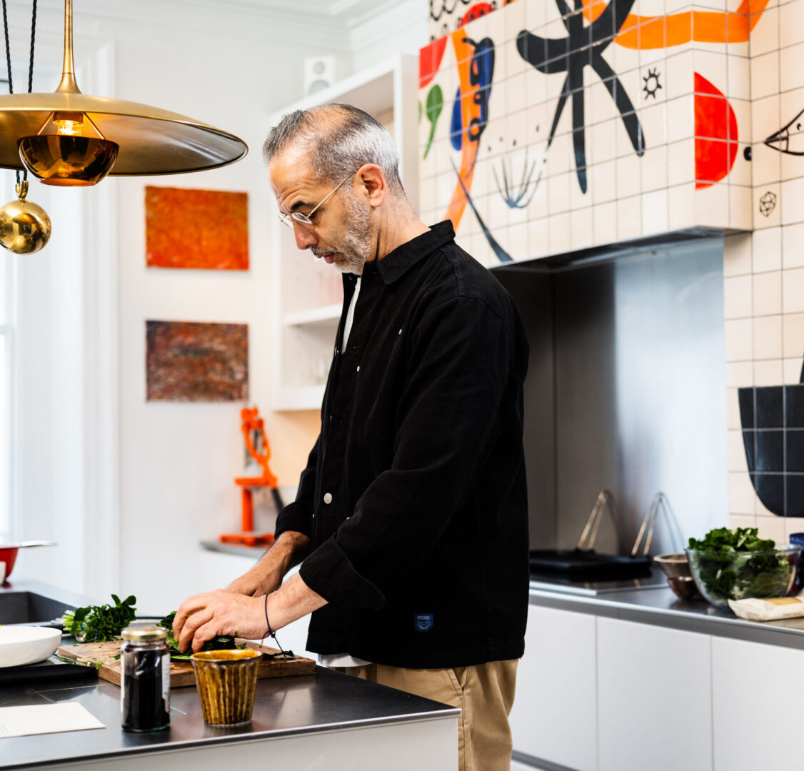 Yotam Ottolenghi preparing food in his Roundhouse kitchen, featuring contemporary cabinetry, stainless steel worktops and a colourful tiled splashback.