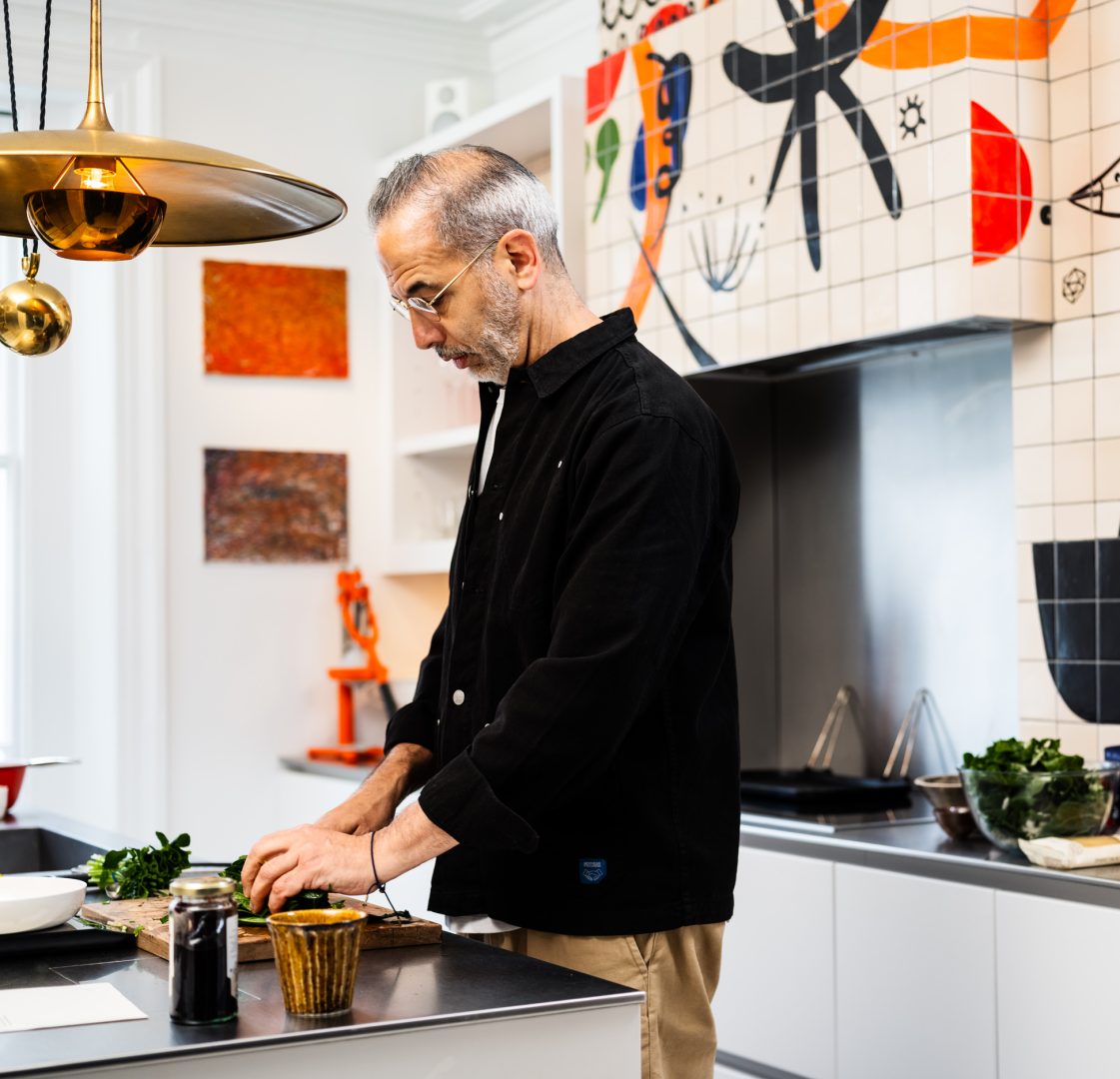 Yotam Ottolenghi preparing food in his Roundhouse kitchen, featuring colourful graphic tiles, white handleless cabinetry and stainless steel worktops.