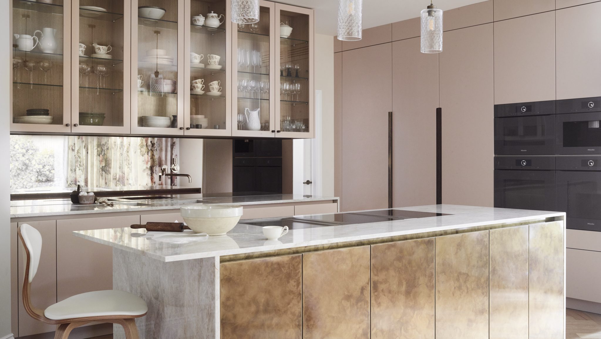Luxury bespoke Roundhouse kitchen featuring soft pink cabinetry, glass-fronted storage, a brass-clad island and natural stone worktops, combining warm colours and metallic finishes.