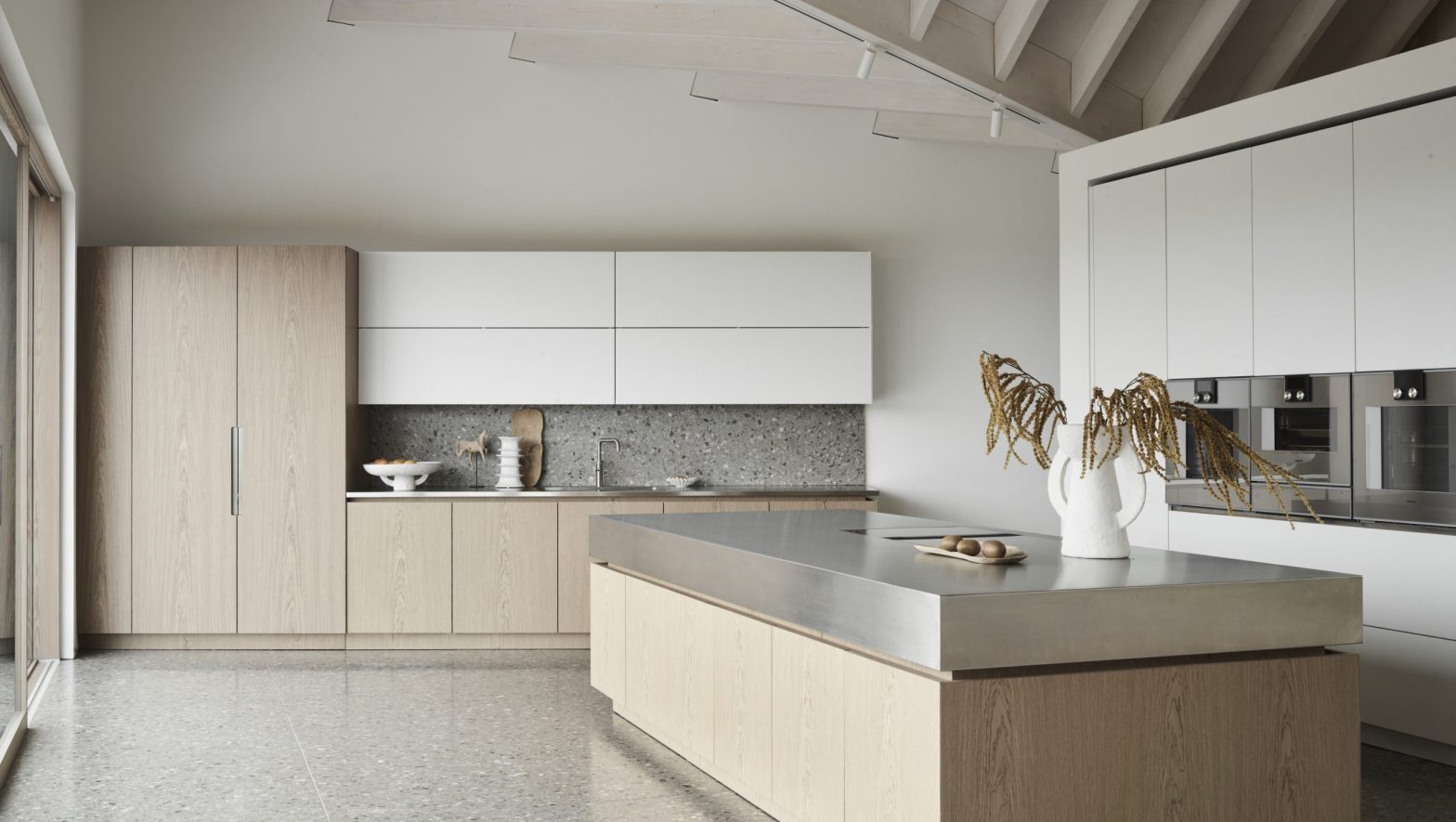 Modern architectural kitchen featuring pale oak cabinetry, a large stone-topped island, integrated appliances and a pitched ceiling with exposed beams.