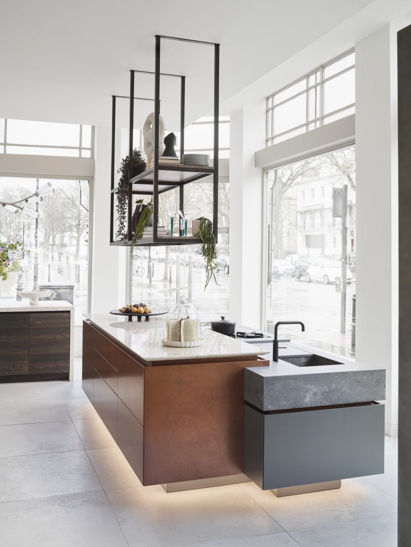 Modern bespoke kitchen with copper-toned island cabinetry, suspended black metal shelving, large windows and warm integrated lighting beneath the island.