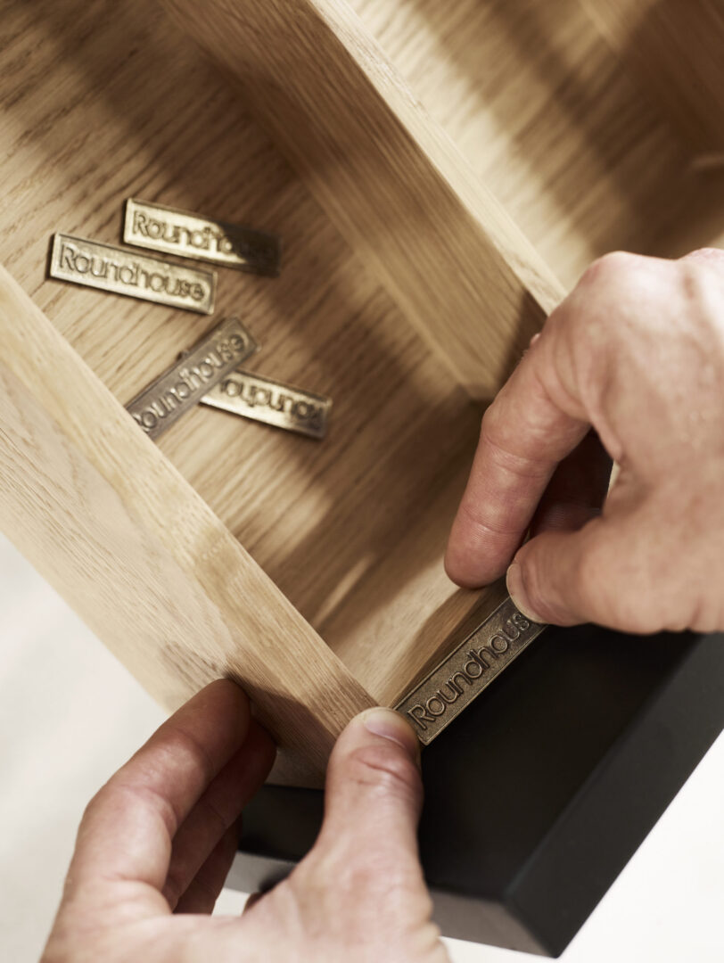 Close-up of a craftsman fitting Roundhouse brass branding to a handcrafted timber drawer, showcasing British kitchen cabinetry craftsmanship.