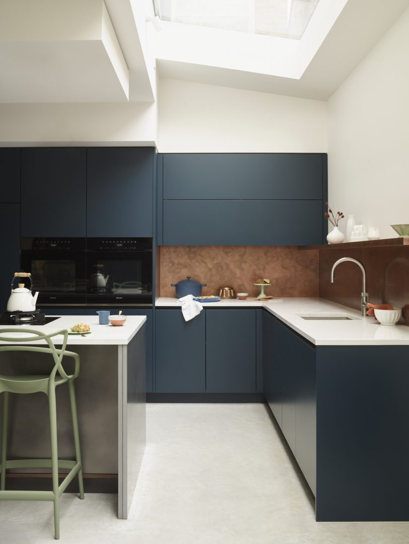 Deep blue bespoke kitchen cabinetry paired with a warm copper splashback, white worktops and skylights, creating contrast between cool and warm tones.