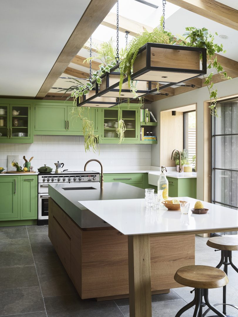 Botanical green kitchen cabinetry with a natural oak island, exposed timber beams and suspended plants, connecting Roundhouse's Oliver design with nature.
