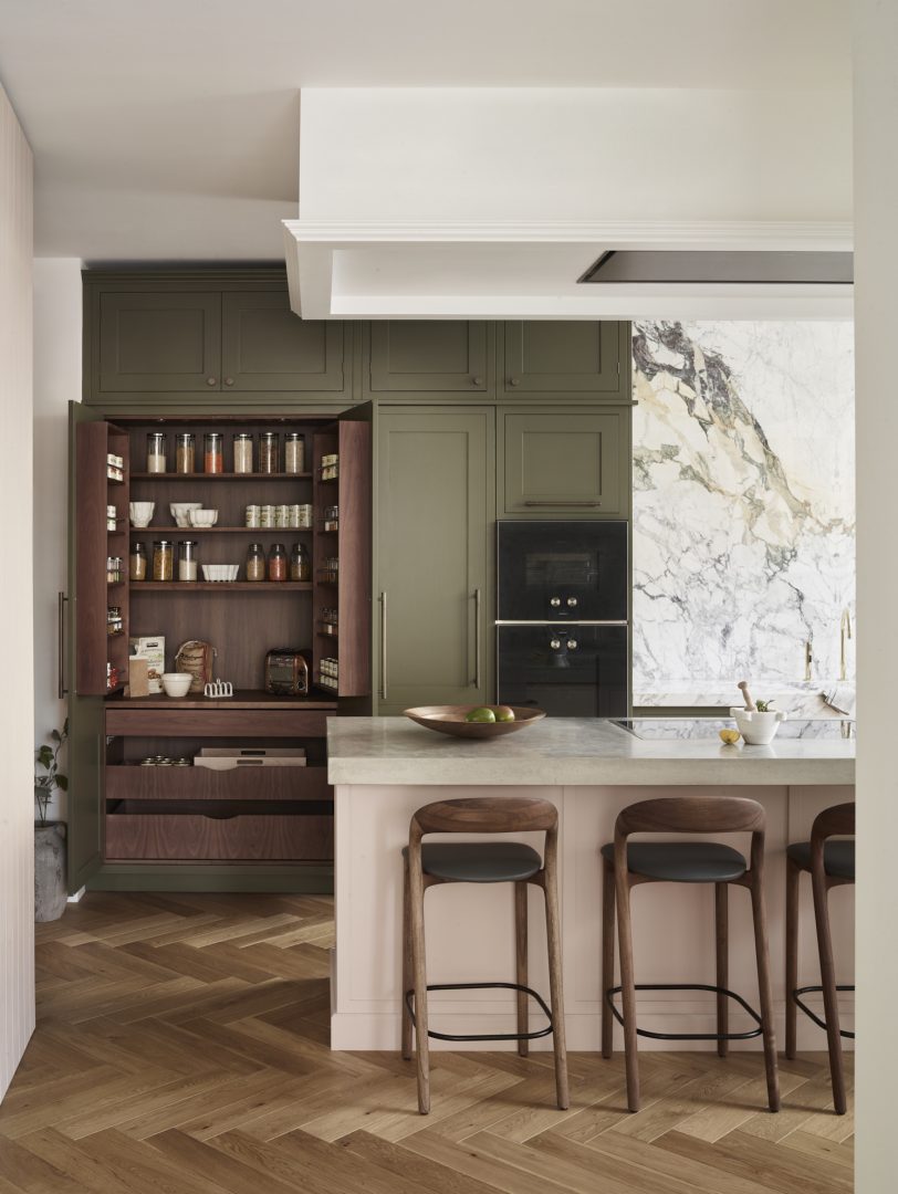 Olive green kitchen cabinetry with an open walnut pantry, marble splashback and soft pink island, combining earthy tones in a bespoke Roundhouse design.